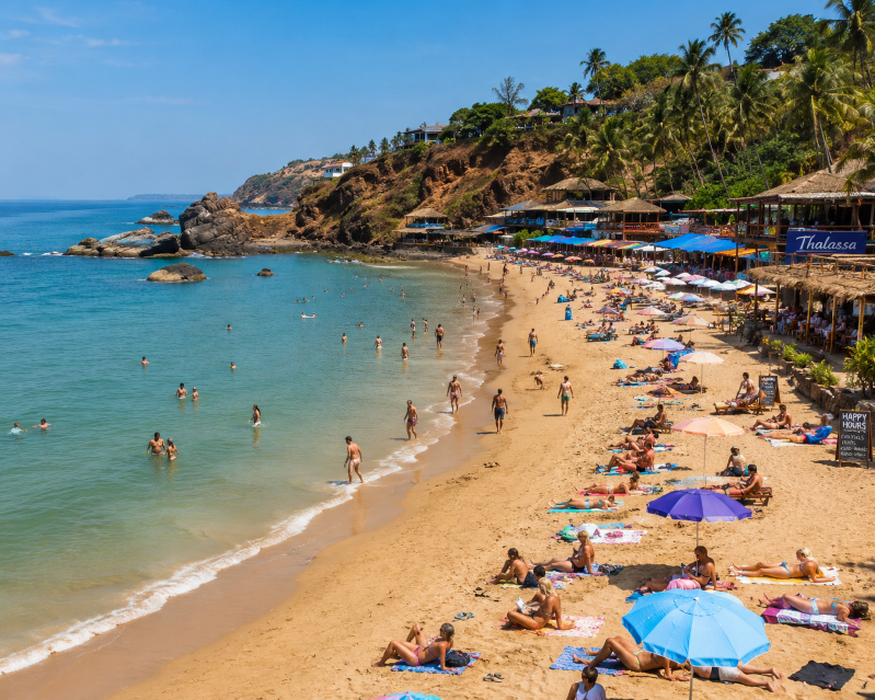 Tourists enjoying swimming, sunbathing, and relaxing at the scenic Vagator Beach in Goa, surrounded by golden sands, turquoise Arabian Sea waters, rocky cliffs, palm trees, and lively beach shacks under a bright tropical sky.