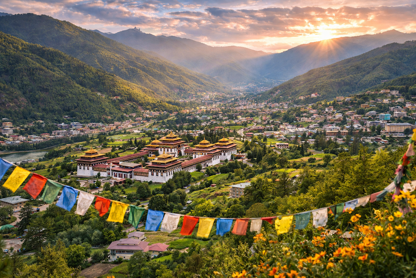 Panoramic sunset view of Thimphu nestled in a lush Himalayan valley, featuring Tashichho Dzong, traditional Bhutanese architecture, colorful prayer flags, and surrounding green mountains.