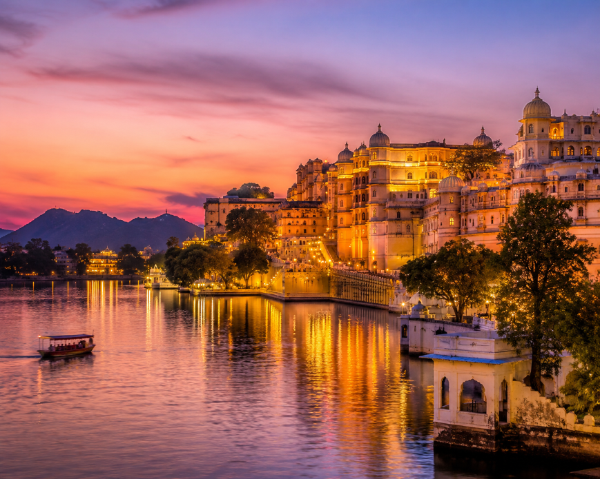 Golden sunset view of the majestic City Palace in Udaipur reflecting over the calm waters of Lake Pichola, with illuminated heritage buildings, Aravalli hills, and a traditional boat creating a romantic royal atmosphere.